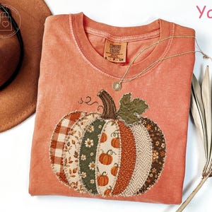 May include: A yam-colored crewneck t-shirt featuring a large pumpkin graphic made of various fabric patches in plaid, floral, and herringbone patterns. The shirt has a Comfort Colors label at the inner neckline and a raw-edge stitched pumpkin design.