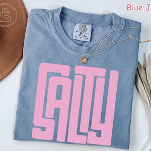 May include: Blue jean colored t-shirt with the word "SALTY" in large pink block letters. The shirt has a crew neck and short sleeves. A gold necklace with a pendant is draped across the shirt.