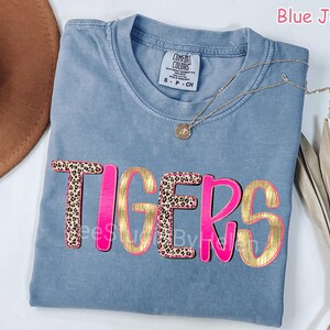 Peut inclure: T-shirt bleu jean Comfort Colors avec le mot "TIGERS" dans un design rose et dor&eacute;. Les lettres "T" et "E" ont un motif l&eacute;opard. Un collier en or avec un pendentif est drap&eacute; sur le t-shirt.