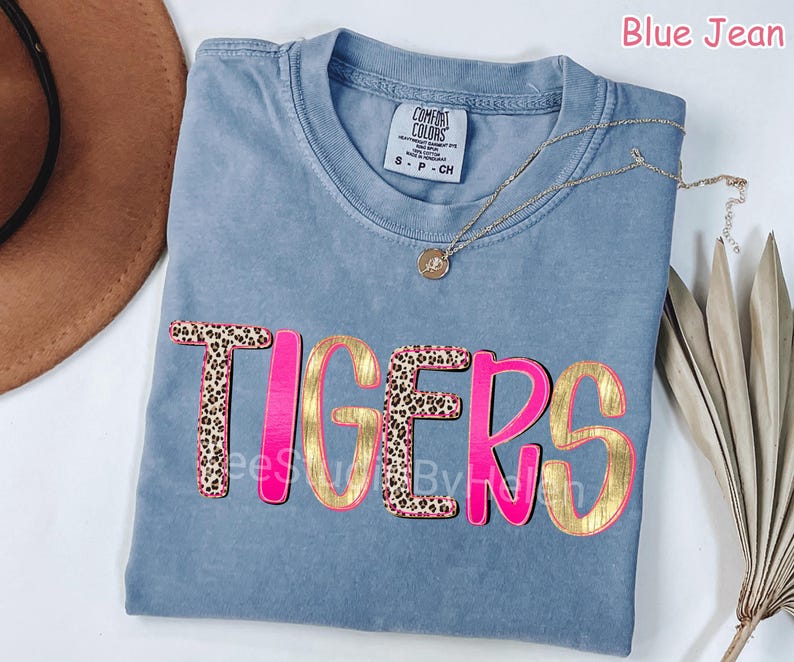 Peut inclure: T-shirt bleu jean Comfort Colors avec le mot "TIGERS" dans un design rose et dor&eacute;. Les lettres "T" et "E" ont un motif l&eacute;opard. Un collier en or avec un pendentif est drap&eacute; sur le t-shirt.