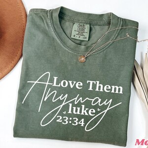 May include: Green t-shirt with white text that reads "Love Them Anyway Luke 23:34".