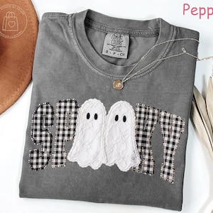 May include: A charcoal gray short-sleeve t-shirt featuring the word SPOOKY in black and white gingham letters, with two white lace ghosts replacing the letter O. The shirt has a crew neckline and a Comfort Colors brand label inside the collar.
