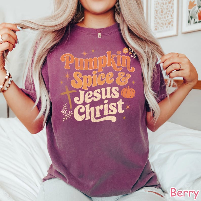Op de afbeelding: Bessenkleurig T-shirt met de tekst "Pumpkin Spice & Jesus Christ" in oranje en cr&egrave;me. Het ontwerp bevat een kruis, een pompoen en bloemaccenten. Het shirt is een casual stijl.