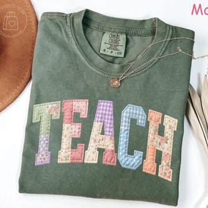 May include: A moss green t-shirt with the word "TEACH" appliqued in colorful patchwork fabric. The shirt has a crew neck and short sleeves. A gold necklace with a pendant rests on the shirt. The shirt is part of a clothing collection.