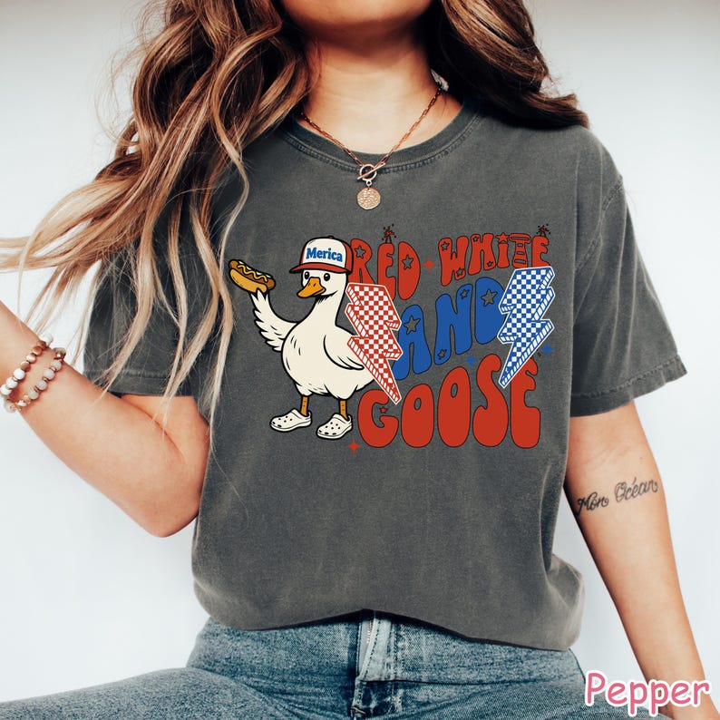 May include: Dark grey t-shirt featuring a cartoon goose wearing a "Merica" hat and holding a hot dog. The shirt has the text "Red White and Goose" in red and blue, with lightning bolt graphics. A fun, patriotic design.