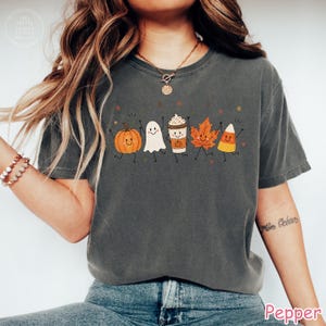 Op de afbeelding: Een antracietgrijs t-shirt met korte mouwen en een grafische print van vijf lachende herfstfiguren: een oranje pompoen, een wit spookje, een koffiebeker, een oranje esdoornblad en een maissnoepje, allemaal met dunne zwarte ledematen.