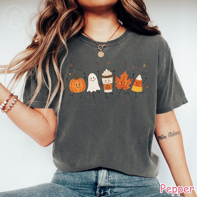 Op de afbeelding: Een antracietgrijs t-shirt met korte mouwen en een grafische print van vijf lachende herfstfiguren: een oranje pompoen, een wit spookje, een koffiebeker, een oranje esdoornblad en een maissnoepje, allemaal met dunne zwarte ledematen.