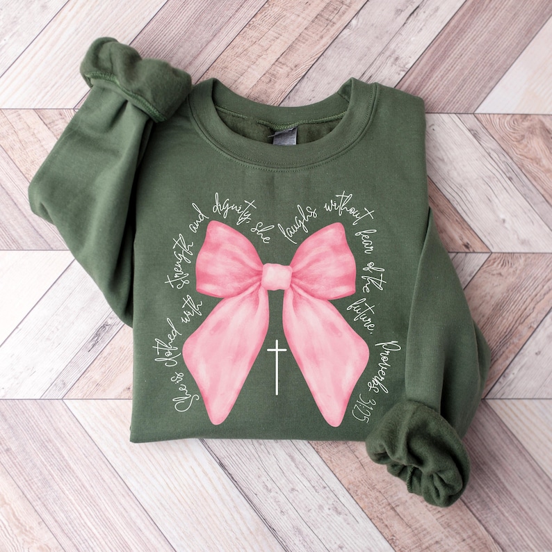 May include: A green sweatshirt with a pink bow and the text "She is clothed with strength and dignity; she laughs without fear of the future. Proverbs 31:25"