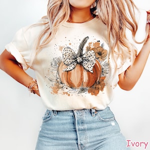 May include: Ivory-colored t-shirt featuring a watercolor-style pumpkin design. The pumpkin is orange with a black and white polka dot bow. Surrounding the pumpkin are floral and leaf illustrations in shades of brown and black.