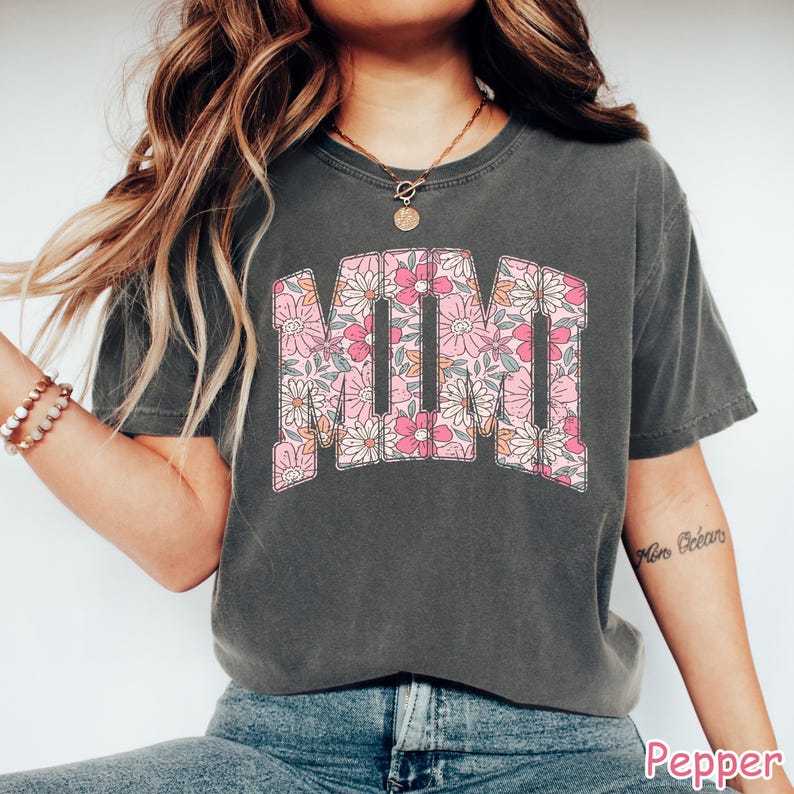 K&ouml;nnte beinhalten: Dunkelgraues T-Shirt mit dem Wort "MOM" in einem floralen Design. Die Buchstaben sind mit rosa, wei&szlig;en und orangefarbenen Blumen gef&uuml;llt. Das Shirt hat kurze &Auml;rmel und einen Rundhalsausschnitt.
