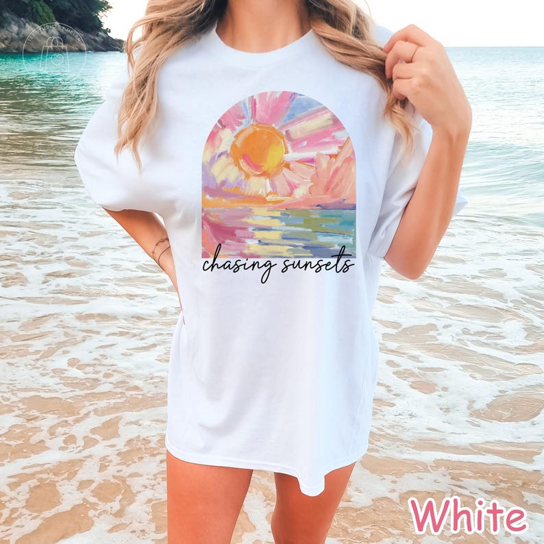 May include: White t-shirt featuring a colorful sunset design within an arch, with the words "chasing sunsets" in a script font. The artwork depicts a vibrant sun, pink and blue sky, and water. The shirt is a casual, short-sleeved style.