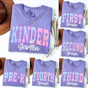May include: A collection of purple crewneck t-shirts featuring block lettering with blue, pink, and floral patterns. The shirts display text including Kindergarten, First Grade, Second Grade, Third Grade, Fourth Grade, and Pre-K in a mix of varsity-style block letters and white cursive script.