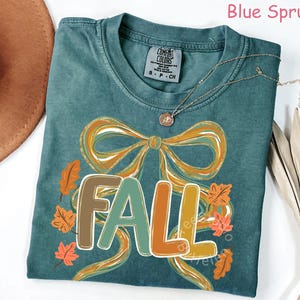 May include: Blue Spruce t-shirt with a fall-themed design. The shirt features the word "FALL" in a colorful, outlined font, with a large bow and autumn leaves. A gold necklace with a pendant is draped on the shirt.