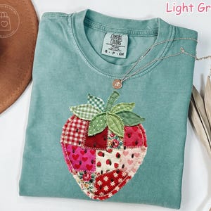 May include: Light green t-shirt with a large, patchwork strawberry design. The strawberry is made of various fabric patterns, including hearts, floral prints, and gingham. A silver necklace with a small pendant rests on the shirt.