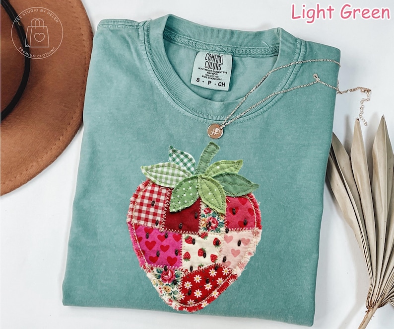 May include: Light green t-shirt with a large, patchwork strawberry design. The strawberry is made of various fabric patterns, including hearts, floral prints, and gingham. A silver necklace with a small pendant rests on the shirt.