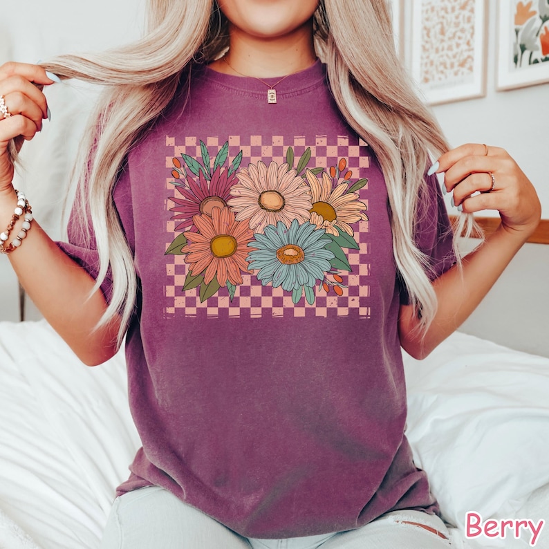 May include: Berry-colored t-shirt featuring a floral design with daisies in shades of pink, orange, and blue, set against a checkered background. The shirt has short sleeves and a relaxed fit, perfect for casual wear.