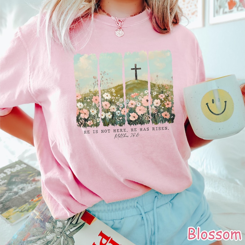 May include: Pink t-shirt with a floral design and a cross on a hill. The text "HE IS NOT HERE. HE HAS RISEN. Matthew 28:6" is printed on the shirt.