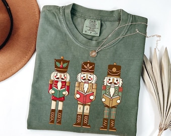 May include: A green t-shirt with a Christmas theme featuring three nutcracker illustrations. The nutcrackers are holding books and are wearing traditional costumes. The text "COMFORT COLORS" is printed on the shirt.
