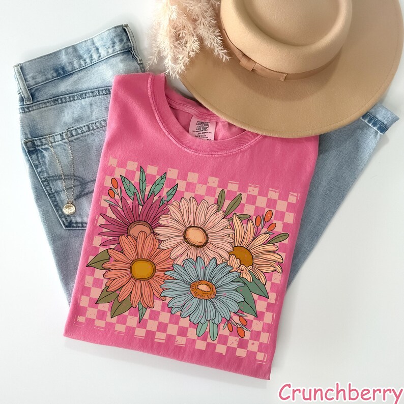 May include: A pink t-shirt with a floral graphic, a tan hat, and light blue jeans. The t-shirt features a checkerboard pattern and a bouquet of colorful flowers. The word "Crunchberry" is printed at the bottom.