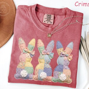 K&ouml;nnte beinhalten: Karmesinrotes T-Shirt mit drei aufgen&auml;hten Patchwork-Hasen. Jeder Hase hat eine Schleife und eine dekorative Blume. Eine silberne Halskette mit Anh&auml;nger befindet sich auf dem T-Shirt. Das T-Shirt hat ein Comfort Colors-Etikett.