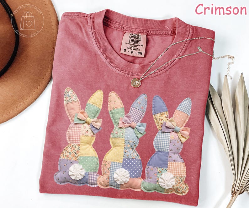 K&ouml;nnte beinhalten: Karmesinrotes T-Shirt mit drei aufgen&auml;hten Patchwork-Hasen. Jeder Hase hat eine Schleife und eine dekorative Blume. Eine silberne Halskette mit Anh&auml;nger befindet sich auf dem T-Shirt. Das T-Shirt hat ein Comfort Colors-Etikett.