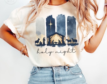May include: Ivory-colored t-shirt featuring a nativity scene with a star, stable, and silhouettes of animals and figures. The words "holy night" are written in a script font below the graphic. The shirt is a relaxed fit.