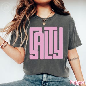 May include: Dark grey t-shirt with the word "SALTY" in large, pink, block letters. The shirt is a crew neck and has short sleeves. The model is wearing blue jeans.
