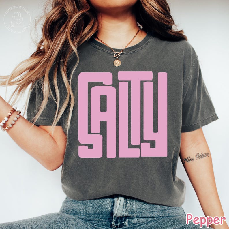 May include: Dark grey t-shirt with the word "SALTY" in large, pink, block letters. The shirt is a crew neck and has short sleeves. The model is wearing blue jeans.