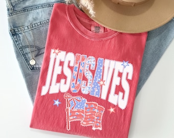 以下が含まれることがあります： 赤と青の模様が入った白い文字「JESUS SAVES」がプリントされたスイカ色のTシャツ。文字の下には、リボンが付いたアメリカ国旗のグラフィックがあります。Tシャツは、水色のジーンズとベージュの帽子と一緒に配置されています。