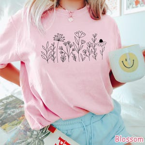 May include: A pink t-shirt with a black line drawing of flowers and leaves. The shirt is worn by a person with light brown hair and blue jeans.
