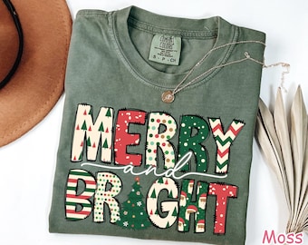 May include: A green t-shirt with the words "Merry and Bright" in colorful, patterned letters. The letters are decorated with Christmas trees, snowflakes, and other festive designs.