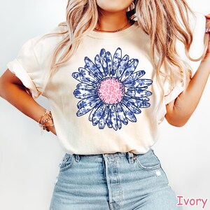 May include: Ivory-colored t-shirt featuring a large blue and white floral design with a pink center. The flower has a detailed pattern. The shirt is paired with blue denim shorts. The word "Ivory" is printed in the bottom right corner.