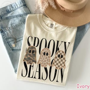 May include: An ivory t-shirt featuring the black text SPOOKY SEASON and three ghost illustrations. The first ghost has a camouflage pattern, the second has a leopard print, and the third has a checkered pattern. The shirt has a crew neckline.