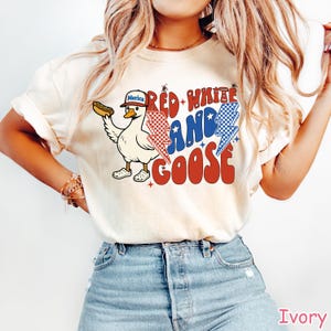 May include: Ivory-colored t-shirt with a graphic of a goose wearing a hat that says "Merica" and holding a hot dog. The text "Red, White, and Goose" is in red, white, and blue. The goose is wearing white clogs.