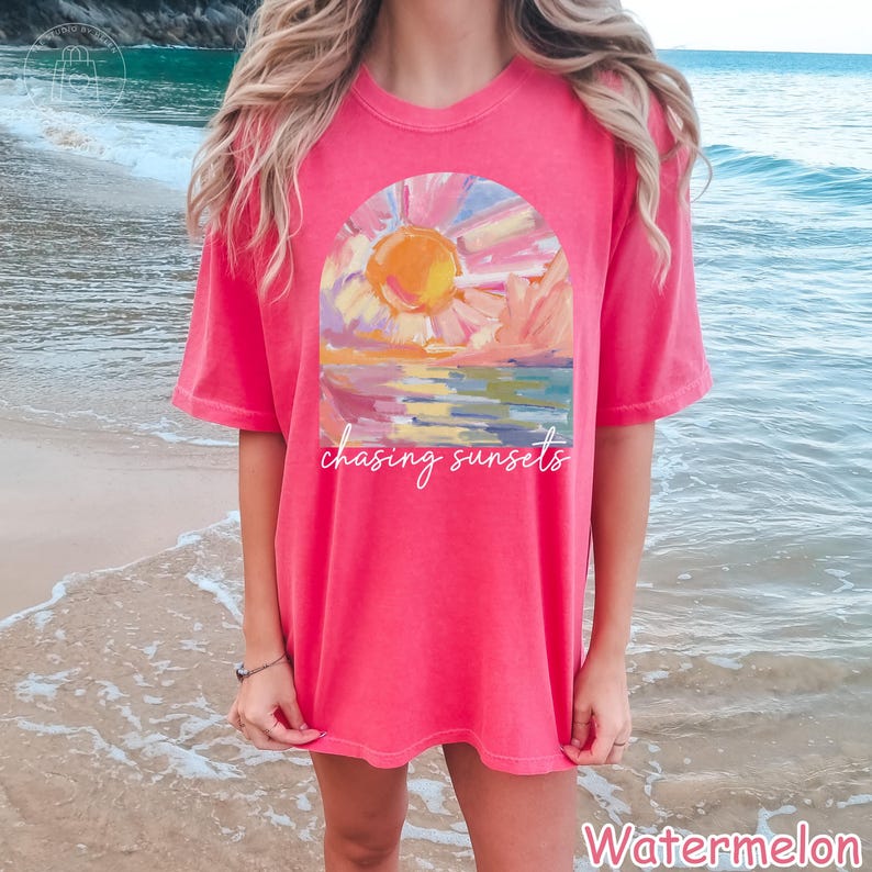 May include: A watermelon-colored t-shirt with a sunset graphic. The graphic features a colorful depiction of a sunset over water, with the words "chasing sunsets" in white script. The shirt is a relaxed fit.