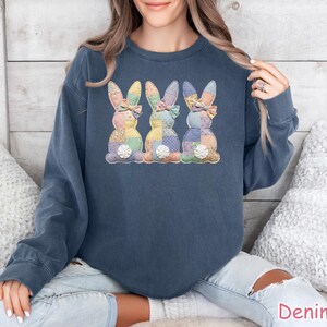 May include: A denim blue sweatshirt featuring three appliqued Easter bunnies. Each bunny has a patchwork design in pastel colors, a bow, and a white flower. The sweatshirt is a casual style.