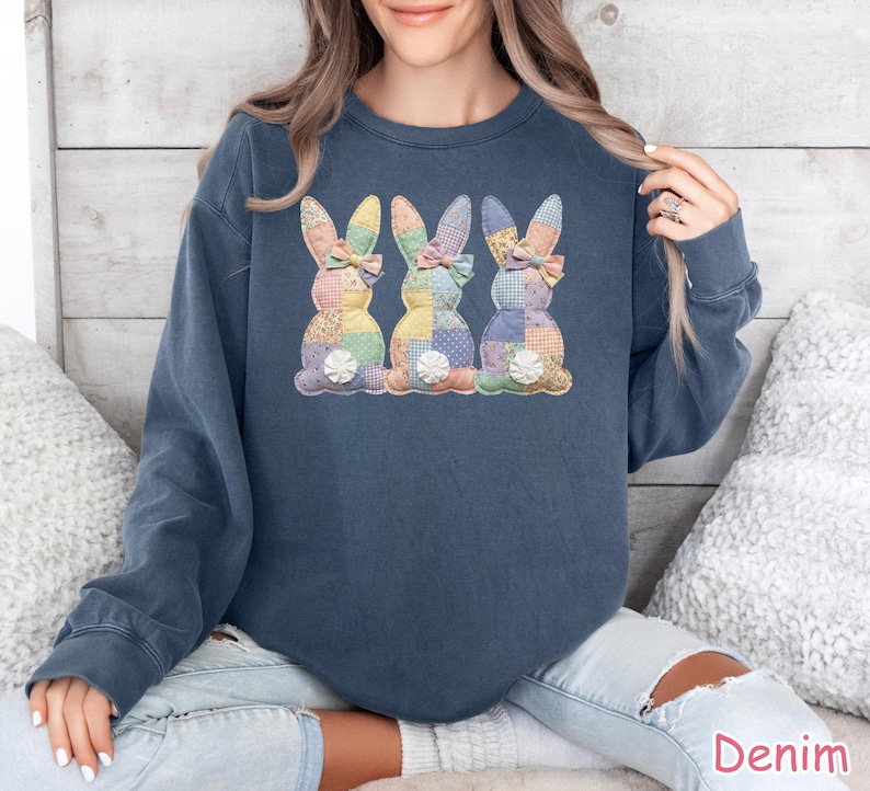 May include: A denim blue sweatshirt featuring three appliqued Easter bunnies. Each bunny has a patchwork design in pastel colors, a bow, and a white flower. The sweatshirt is a casual style.