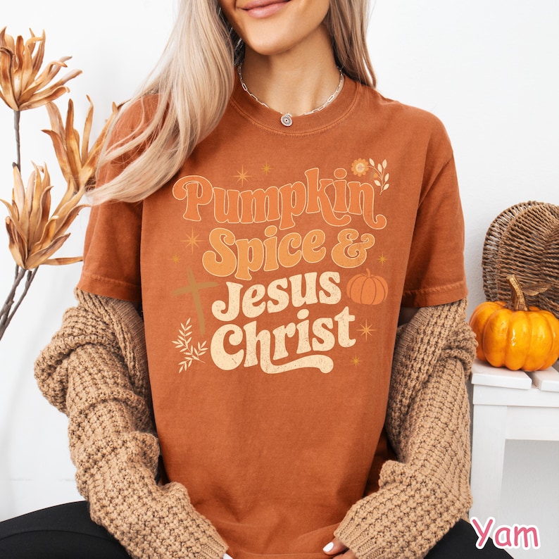 Op de afbeelding: Roestkleurig T-shirt met de tekst "Pumpkin Spice & Jesus Christ" in oranje en cr&egrave;me. Het ontwerp bevat een kruis, een pompoen en bloemaccenten. Het shirt wordt gedragen met een bruin vest.