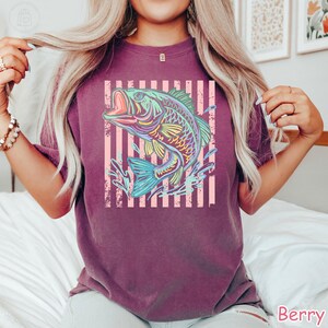 May include: A berry-coloured short-sleeve t-shirt featuring a graphic print of a jumping bass fish in vibrant neon colours. The fish is centred over a background of vertical light pink stripes, creating a bold contrast against the dark fabric.