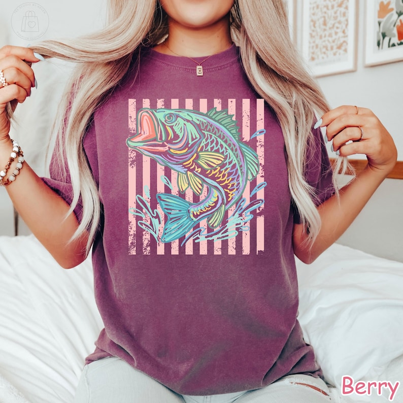 May include: A berry-coloured short-sleeve t-shirt featuring a graphic print of a jumping bass fish in vibrant neon colours. The fish is centred over a background of vertical light pink stripes, creating a bold contrast against the dark fabric.