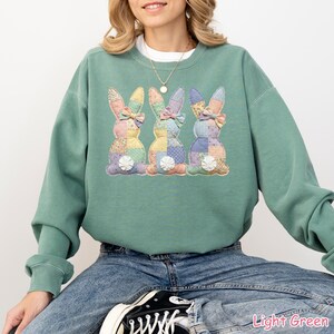May include: Light green sweatshirt featuring three appliqued Easter bunny designs. Each bunny has a patchwork body and a bow. The sweatshirt is a relaxed fit and has long sleeves. The text "Light Green" is at the bottom right.