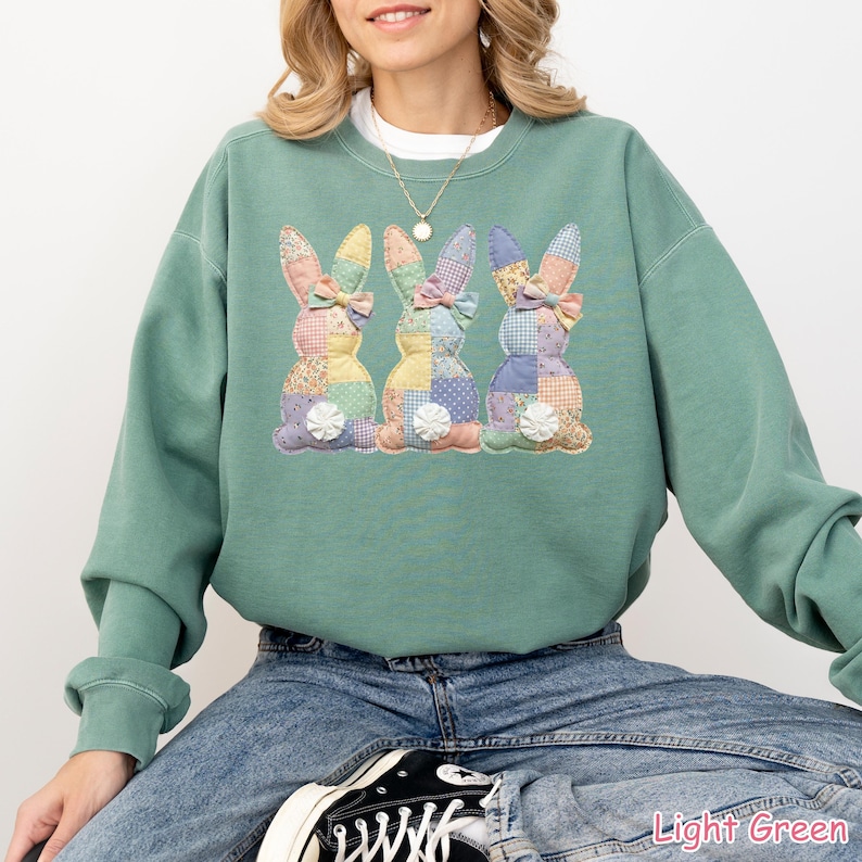 May include: Light green sweatshirt featuring three appliqued Easter bunny designs. Each bunny has a patchwork body and a bow. The sweatshirt is a relaxed fit and has long sleeves. The text "Light Green" is at the bottom right.