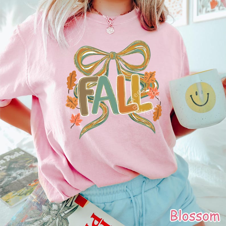 May include: A light pink t-shirt with the word "FALL" in a colorful design. The design includes a large bow, autumn leaves, and the word "FALL" in green, orange, and brown. A white mug with a yellow smiley face is held in the image.