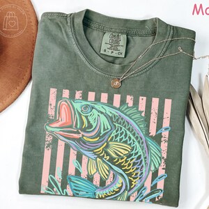 May include: A moss green short-sleeve t-shirt featuring a graphic of a jumping fish in vibrant colours of blue, yellow, and purple. The design includes vertical light pink stripes behind the fish. The shirt has a Comfort Colors brand label.