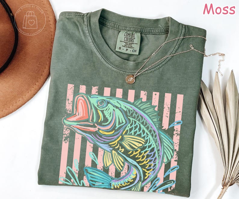 May include: A moss green short-sleeve t-shirt featuring a graphic of a jumping fish in vibrant colours of blue, yellow, and purple. The design includes vertical light pink stripes behind the fish. The shirt has a Comfort Colors brand label.