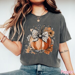 May include: A dark gray t-shirt featuring a watercolor-style pumpkin design. The pumpkin is orange with a polka-dot bow and surrounded by floral accents in shades of orange and brown. The word "Pepper" is printed at the bottom.
