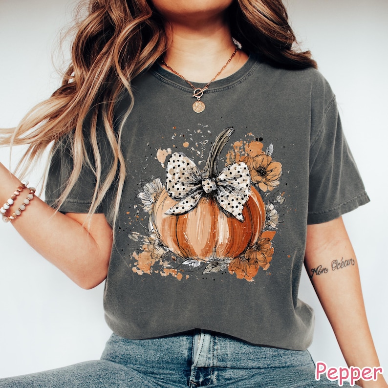 May include: A dark gray t-shirt featuring a watercolor-style pumpkin design. The pumpkin is orange with a polka-dot bow and surrounded by floral accents in shades of orange and brown. The word "Pepper" is printed at the bottom.