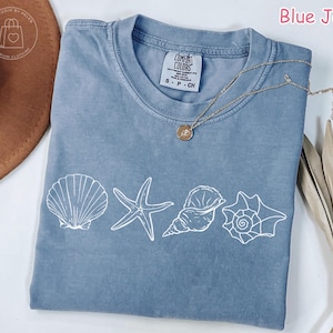 May include: A blue jean-colored t-shirt with a crew neck and short sleeves. The shirt features a white graphic of a seashell, starfish, and two different conch shells. A gold necklace with a pendant is draped across the shirt.