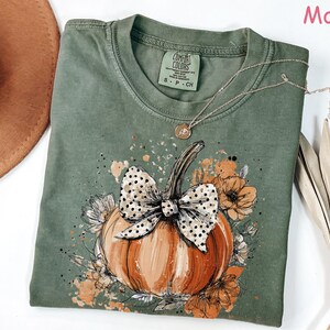 May include: Moss green t-shirt featuring a pumpkin design with a polka dot bow and floral accents. The pumpkin is orange and brown, with a black outline. A gold necklace is draped on the shirt.