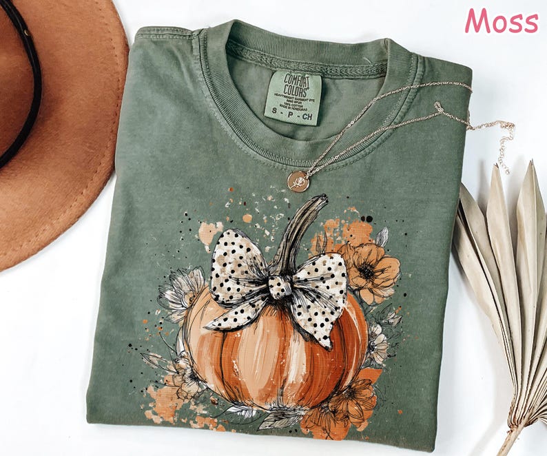 May include: Moss green t-shirt featuring a pumpkin design with a polka dot bow and floral accents. The pumpkin is orange and brown, with a black outline. A gold necklace is draped on the shirt.